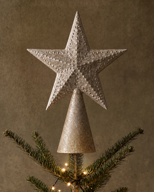 A metallic silver star tree topper mounted on a cone-shaped finial, displayed on top of a decorated Christmas tree with warm lights against a textured brown background.