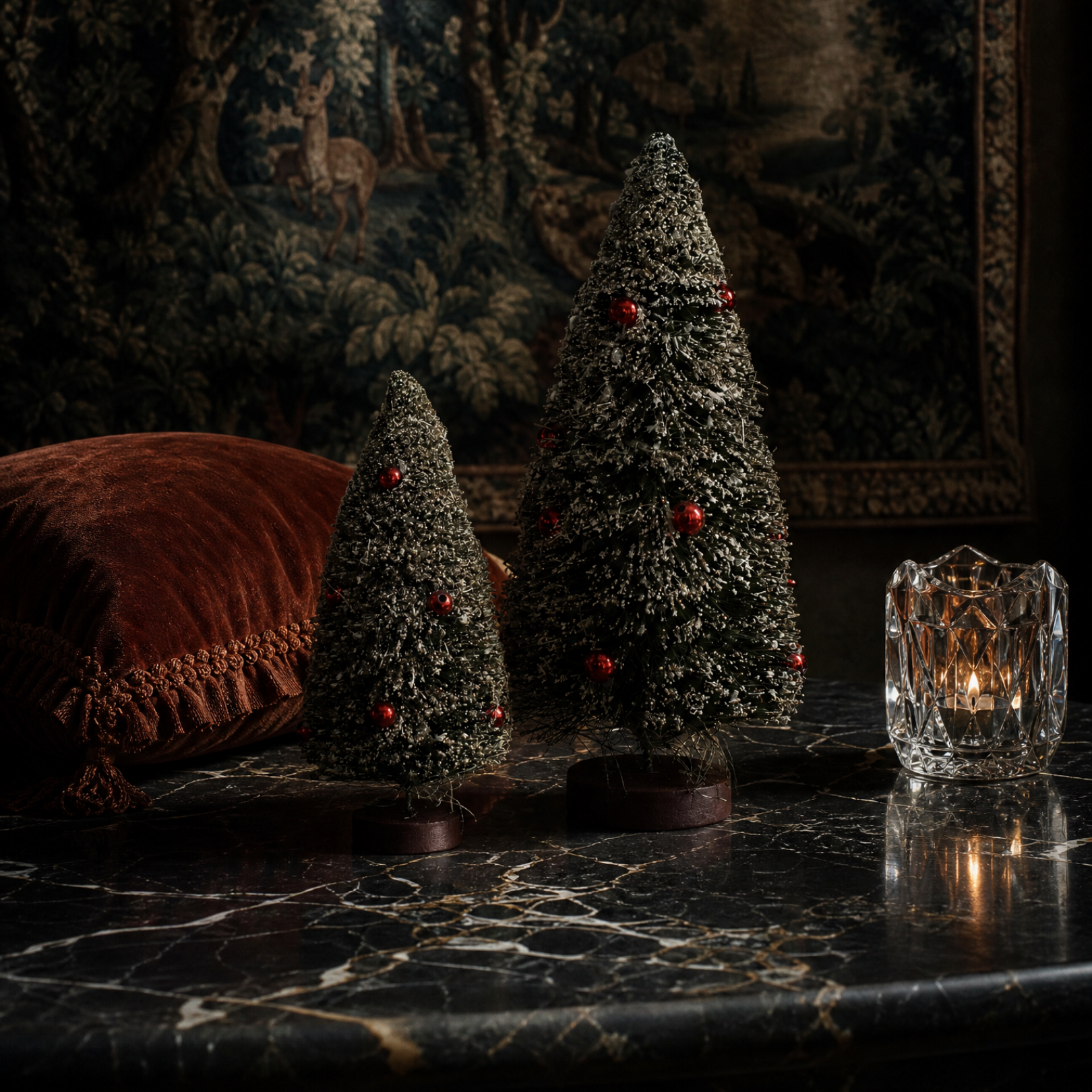Two small frosted Christmas tree ornaments with red berry accents displayed on a dark marble surface, with a lit glass candle holder and a brown velvet pillow in a moody interior setting.