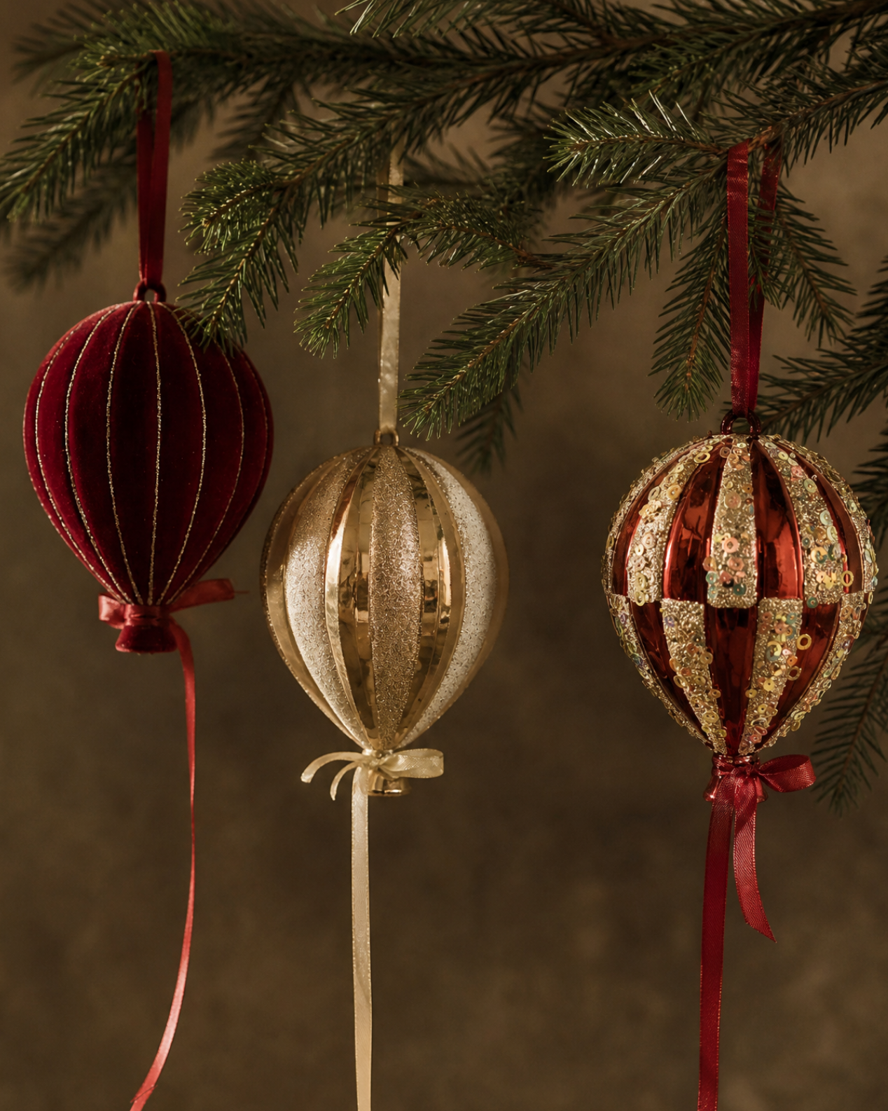 Three decorative Christmas ornaments hang from a pine branch against a dark brown background. The ornaments include a deep red ribbed ball with a long red ribbon, a metallic gold ribbed ball with a cream ribbon, and a red and gold checkered ball with a red ribbon.