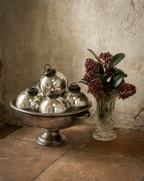 A decorative arrangement of five mirrored silver Christmas ornaments in a silver pedestal bowl, placed beside a clear glass vase with dark red berries and green leaves, set against a rustic textured beige wall and stone surface.