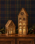 Two decorative house-shaped lanterns with warm glowing lights, displayed on a wooden cabinet against a dark blue plaid wallpaper background with holiday greenery and red berries.