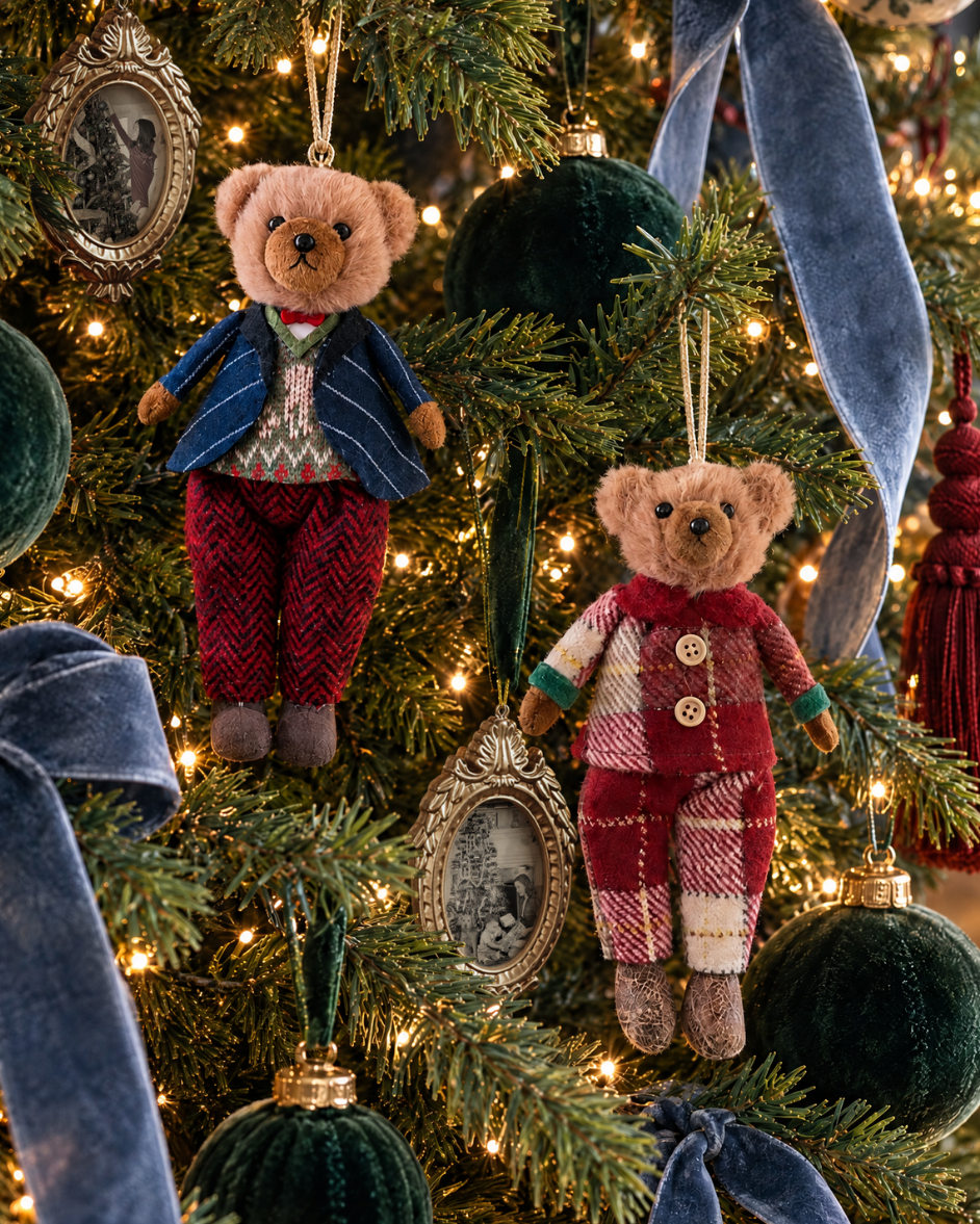Two teddy bear Christmas ornaments hanging on a decorated Christmas tree. One bear wears a blue jacket and red patterned pants, and the other wears a red patterned jacket and cream patterned pants.