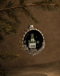 A round Christmas ornament hanging from a pine branch against a warm brown background. The ornament has a scalloped silver rim, a black center with white snowflakes, and a small white house with green tree details.