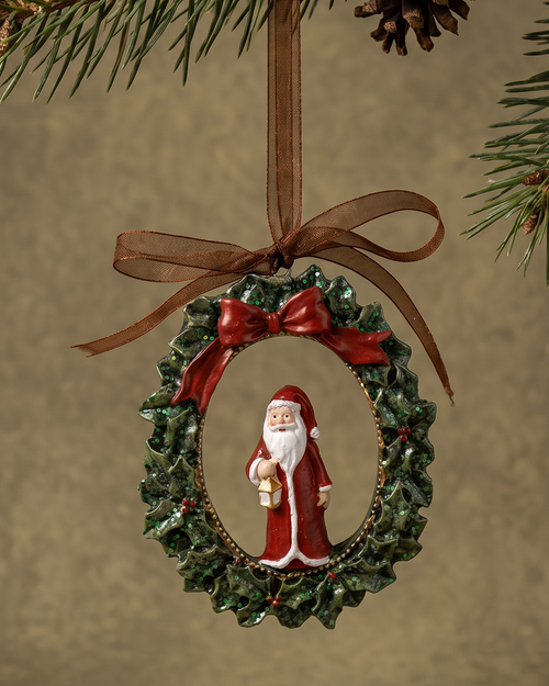 A Christmas ornament shaped like a green wreath with a red bow and a small Santa Claus figure in the center, hanging from a brown ribbon under evergreen tree branches against a warm beige background.