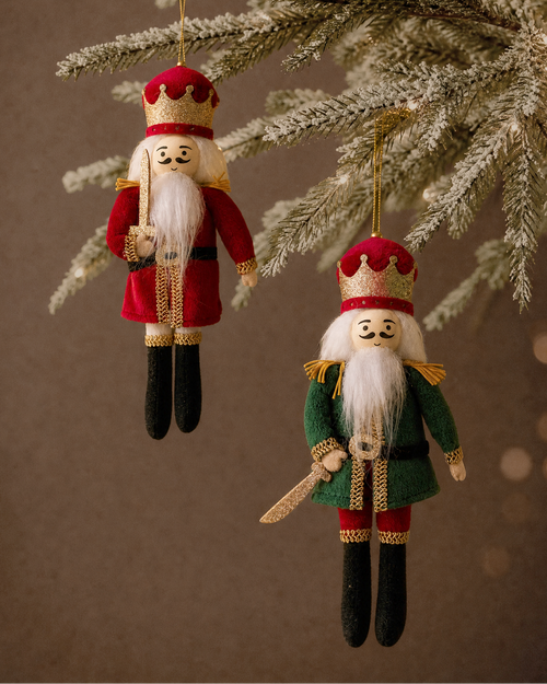 Two hanging Christmas ornament figurines shaped like festive characters, one in red and one in green, suspended from a frosted evergreen branch against a warm brown background.
