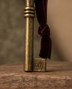 An antique-style brass key with a dark burgundy velvet ribbon tied to its shank, resting on a rustic wooden surface against a warm, blurred background. The key features the monogram initials 'CD' on its bow. Magic Santa Key "Claus Delivery" Engraving