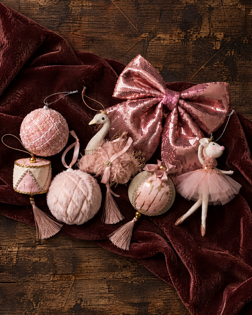 A set of pink and blush ballet-themed Christmas ornaments arranged on a dark burgundy velvet cloth over a rustic dark wood background. The ornaments include a large sequined bow, a ballerina figure, a swan, and textured ball ornaments.