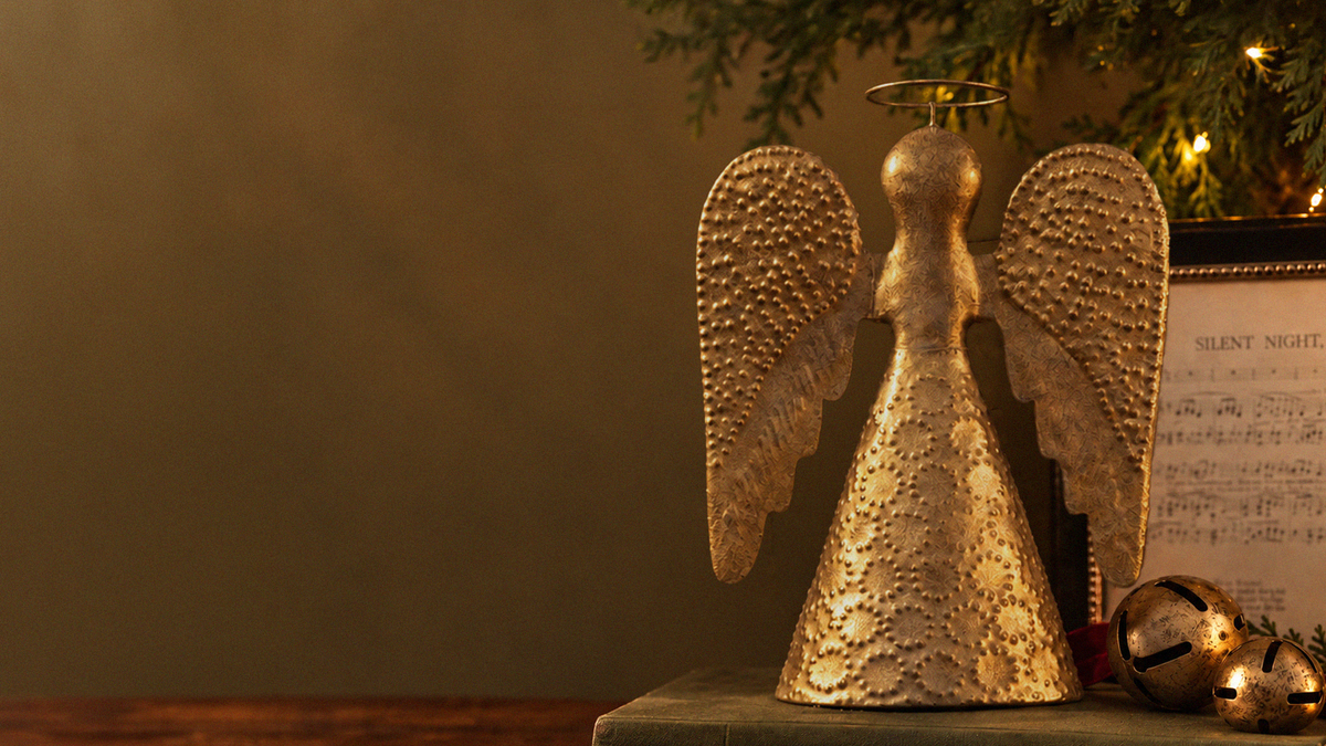 A gold angel figurine with textured wings and a halo stands on a tabletop beside a framed sheet music print and small metallic bells, with warm holiday lighting in the background.