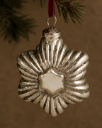 A silver metallic snowflake Christmas ornament with a textured, ribbed outer design and a central white star, hanging from a red ribbon on a pine tree branch against a warm brown background.
