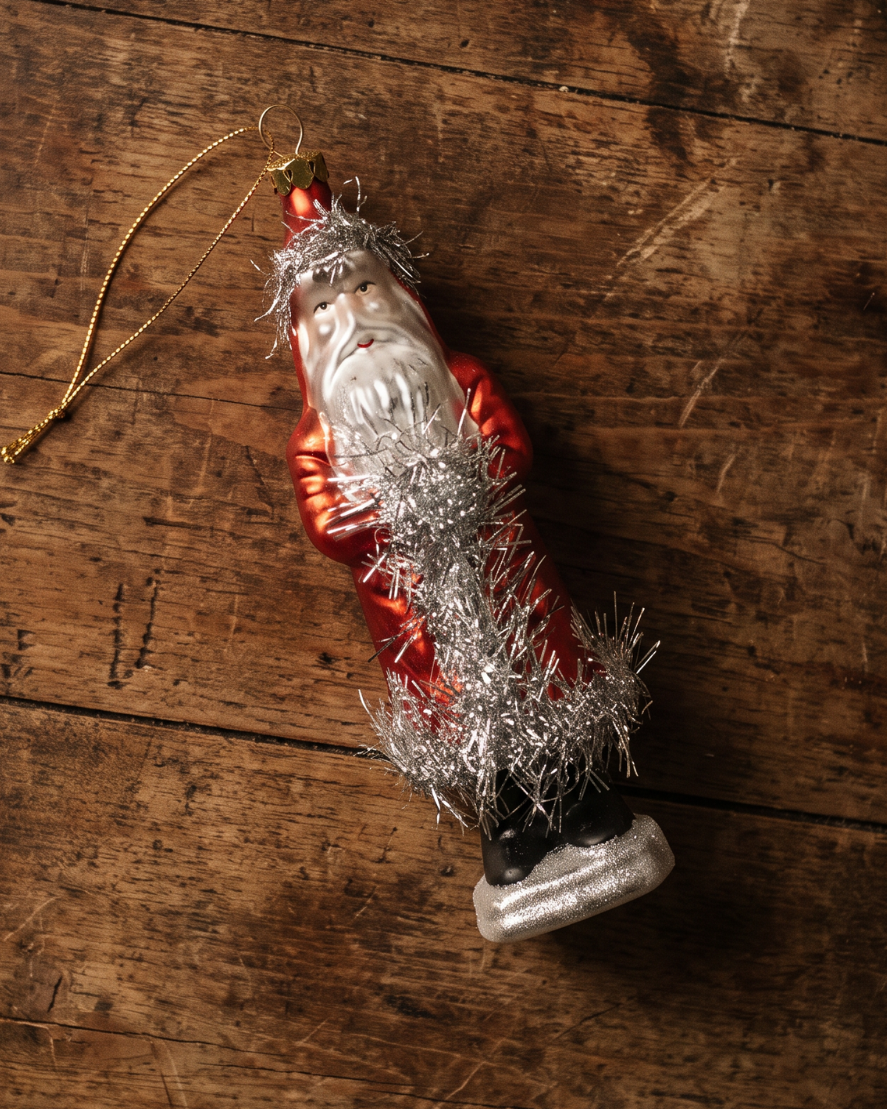 A vintage-style Christmas ornament shaped like Santa Claus, featuring a shiny red suit, silver tinsel, and a gold hanging string, displayed diagonally on a rustic dark wood surface.