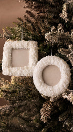 Two white textured Christmas ornaments, one square and one round wreath-shaped, hanging on a frosted evergreen tree against a warm brown background.