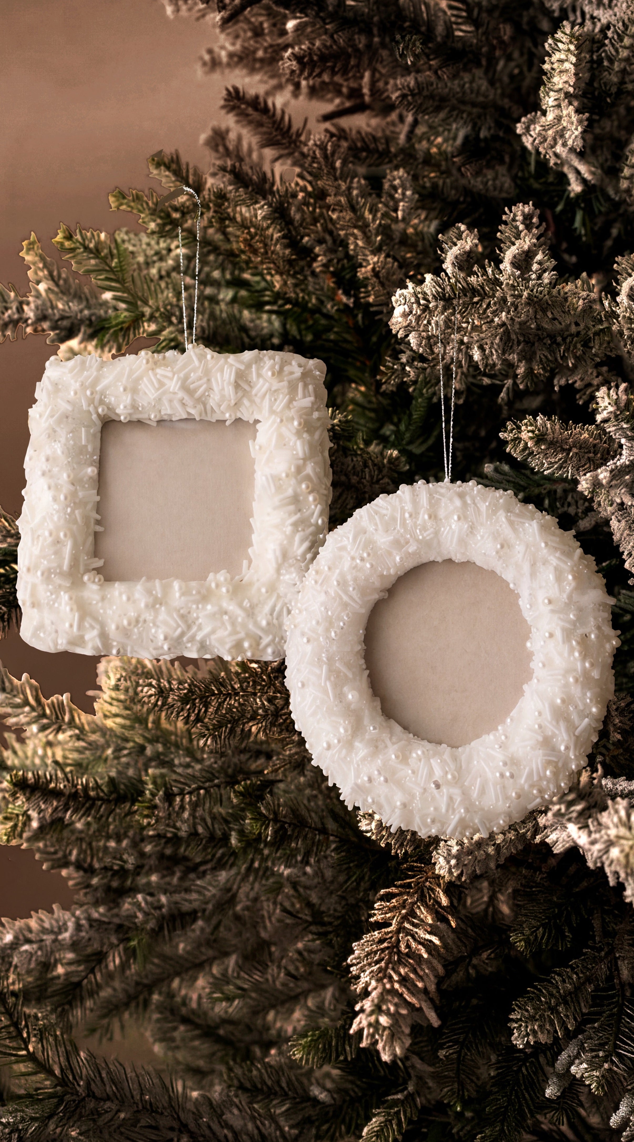 Two white textured Christmas ornaments, one square and one round wreath-shaped, hanging on a frosted evergreen tree against a warm brown background.