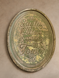 An oval vintage-style metal sign with distressed green and gold patina, featuring the text 'Santa Claus is coming to town' and numbers 12 and 25, mounted on a textured beige plaster wall.