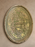 An oval vintage-style metal sign with distressed green and gold patina, featuring the text 'Santa Claus is coming to town' and numbers 12 and 25, mounted on a textured beige plaster wall.