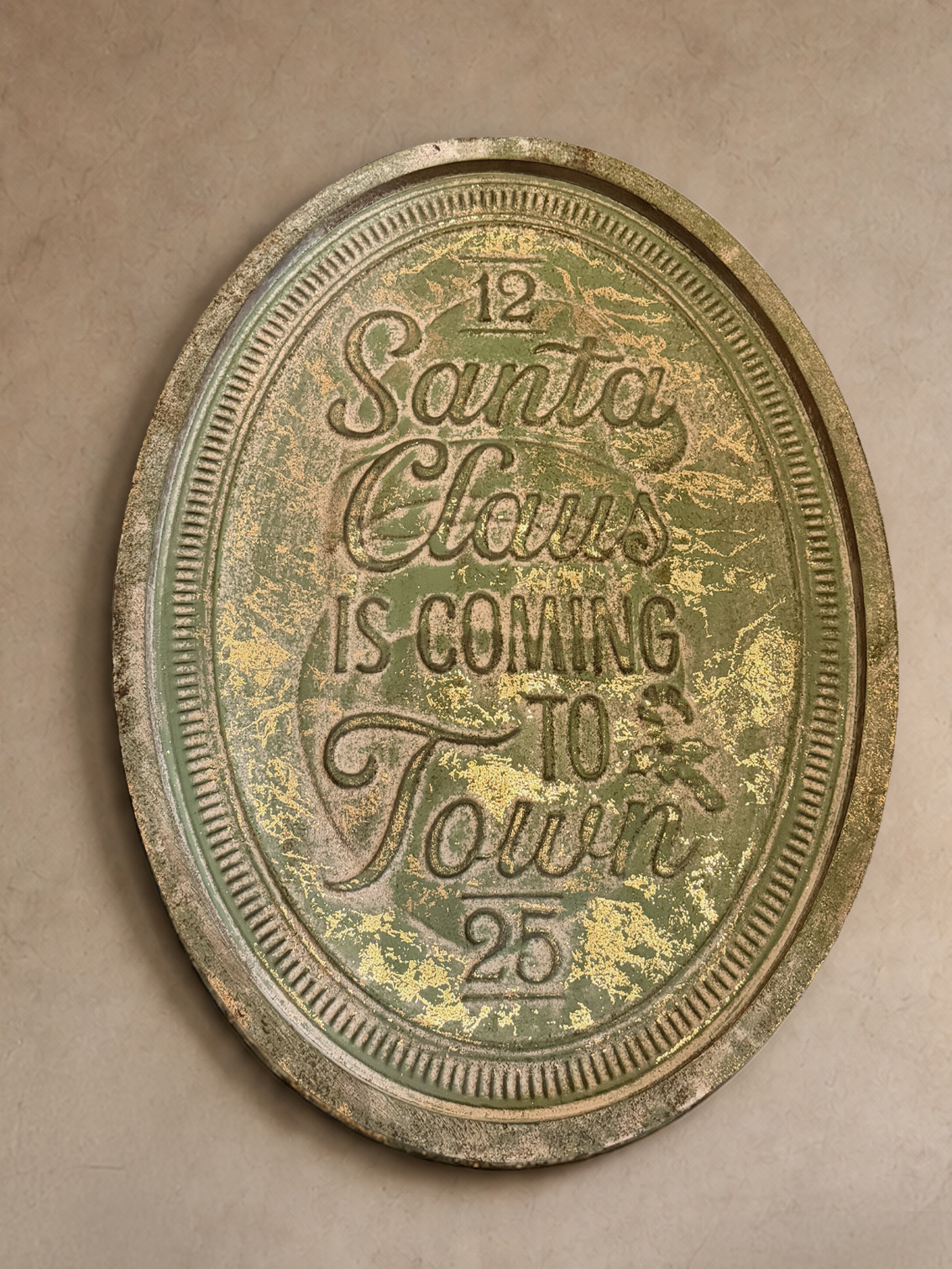 An oval vintage-style metal sign with distressed green and gold patina, featuring the text 'Santa Claus is coming to town' and numbers 12 and 25, mounted on a textured beige plaster wall.